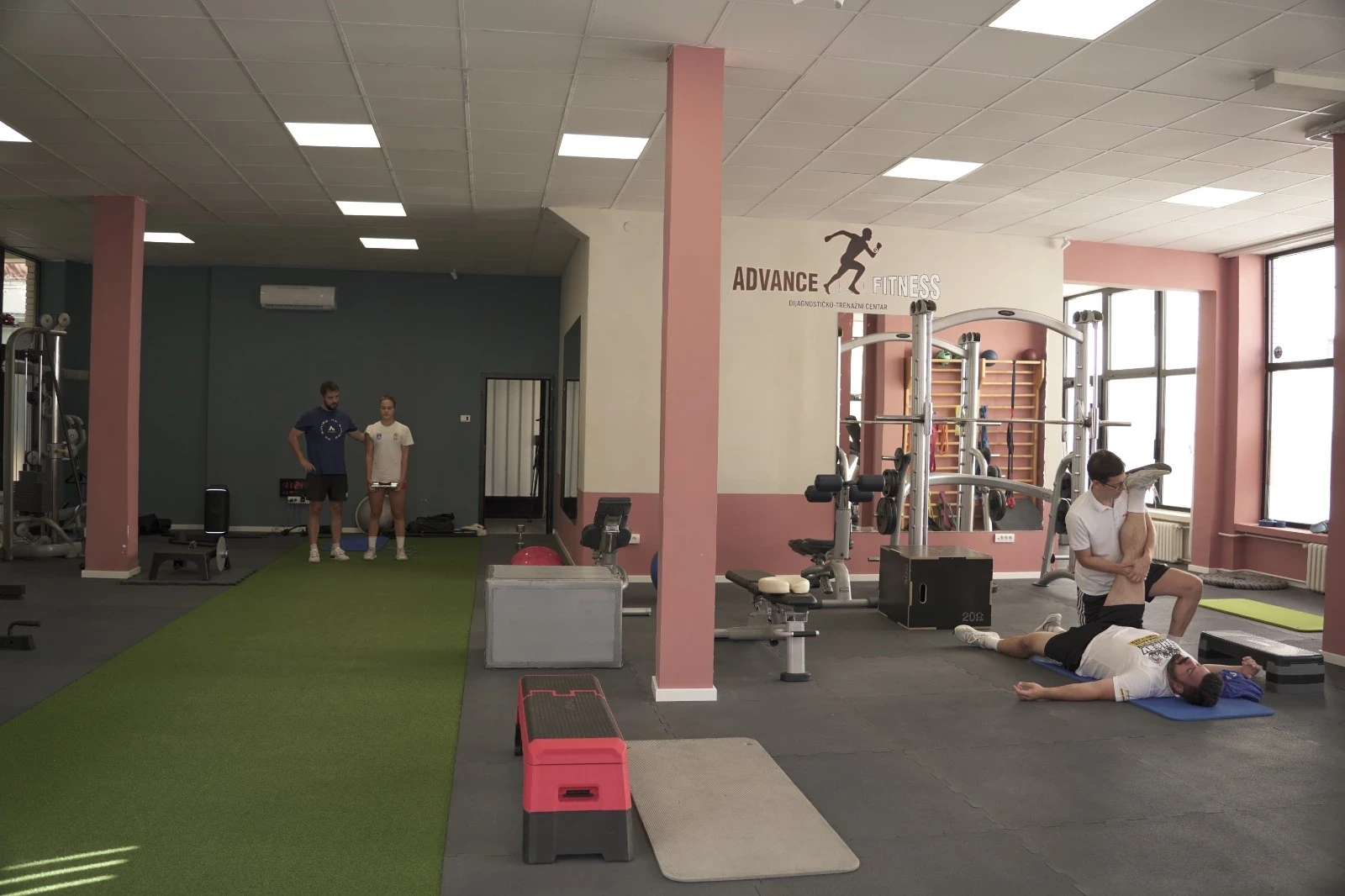 Pogled na ceo prostor centra — levo staza od veštačke trave na kojoj trener vodi sportistkinju, desno deo sale ispod znaka centra sa radom na strunjači.