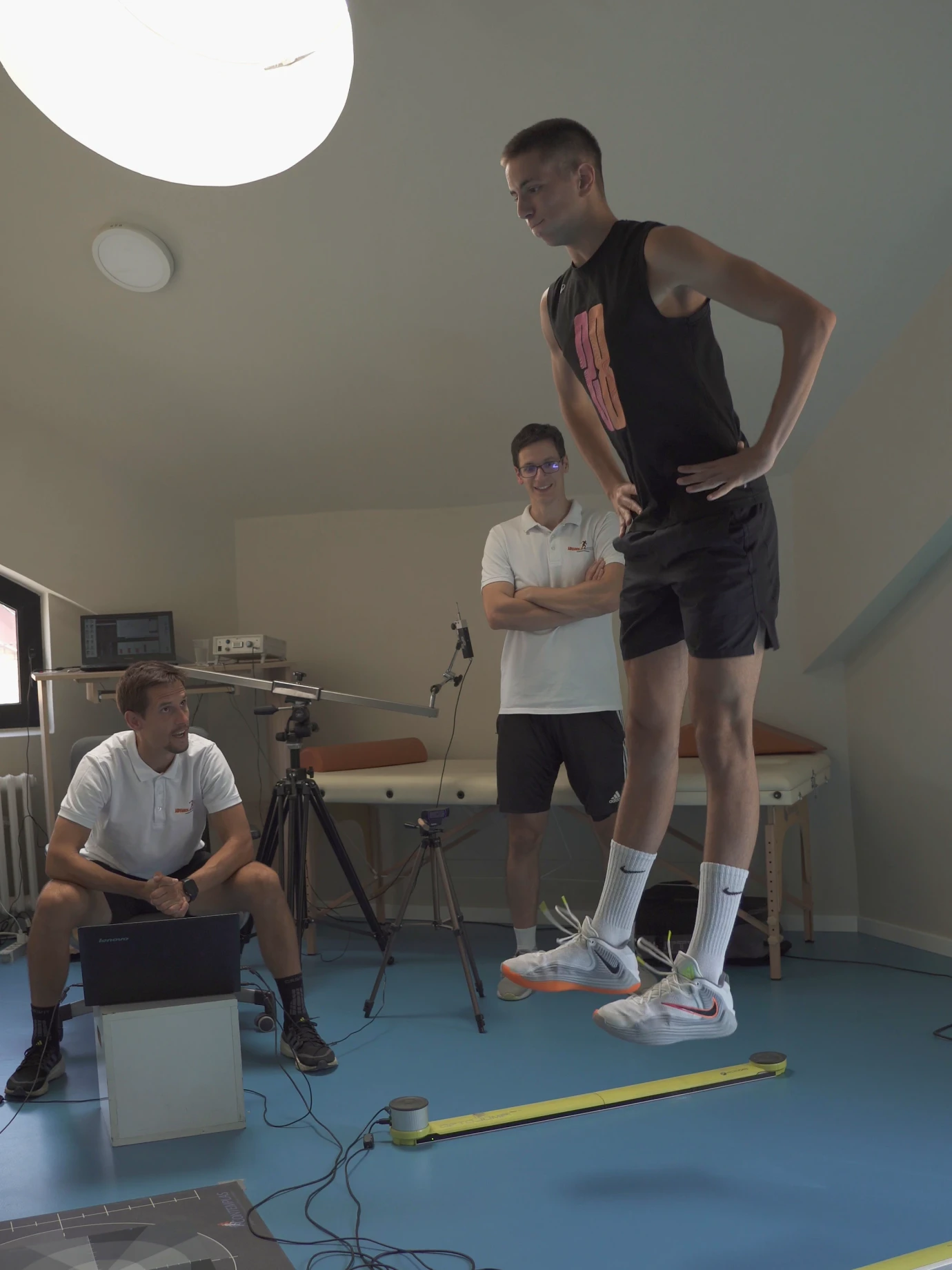 Mladi sportista stoji između Optojump letvi u prostoriji za merenje. Jedan trener sedi za laptopom i prati merenje, drugi stoji pored njega.