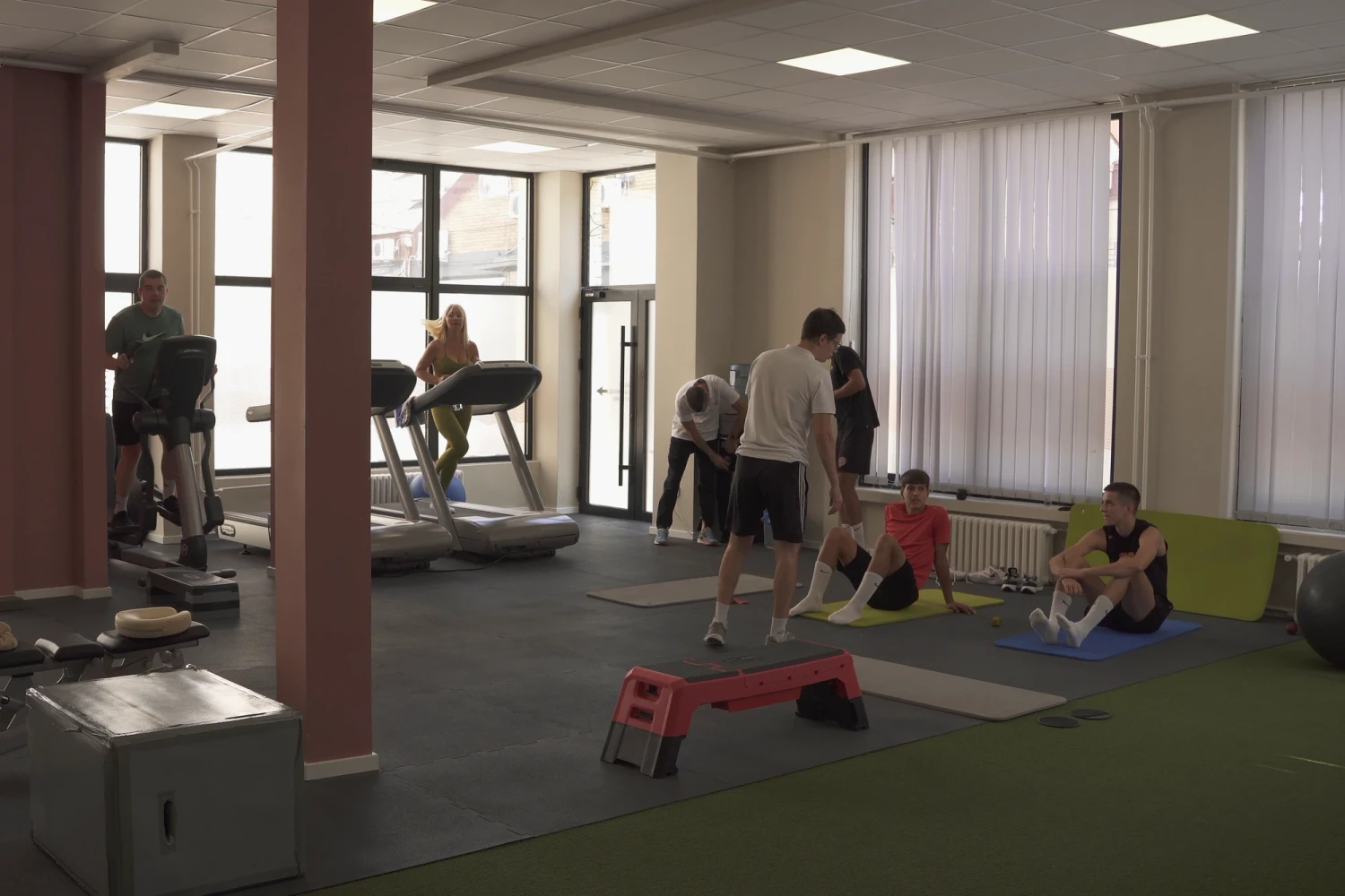 Kardio zona centra — trake za trčanje i eliptični trenažer uz prozore, a u desnom delu sale sportisti rade na strunjačama.