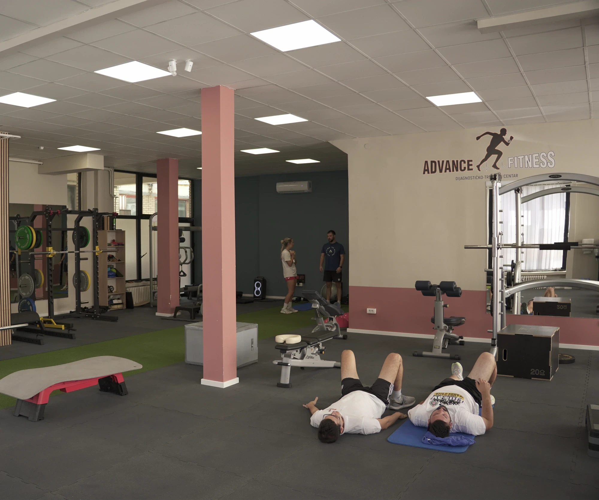 Sala Advance Fitness Centra na Novom Beogradu — stalci za tegove, prostor za rad na podu i znak centra na zidu.