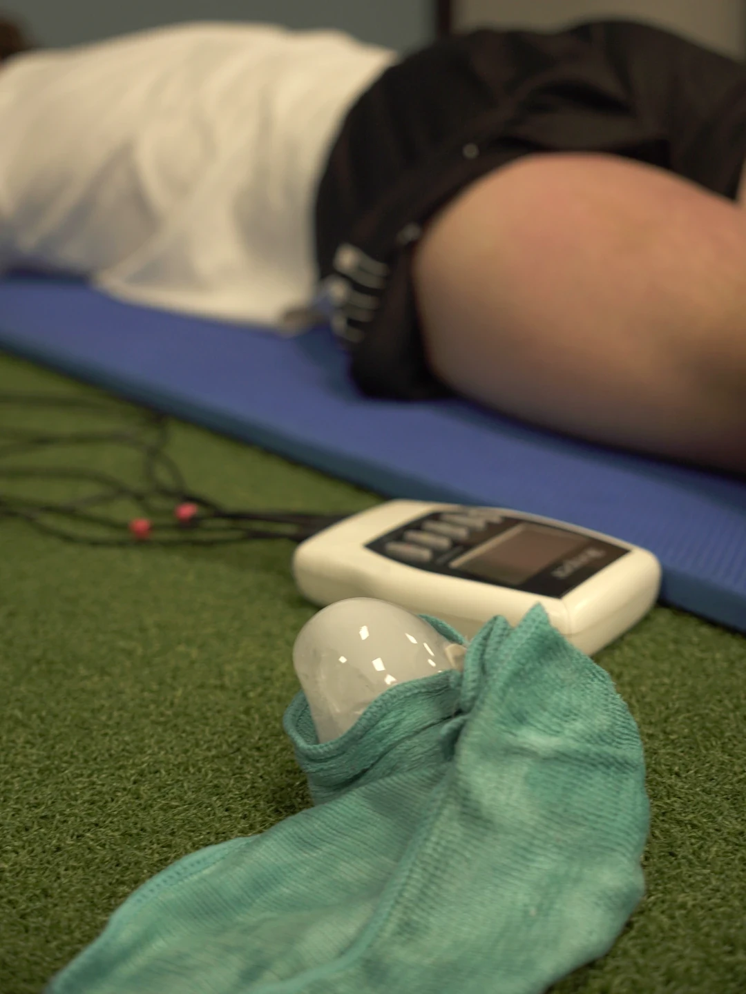 Compex za elektrostimulaciju i led u krpi na podu, pored sportiste koji leži na strunjači sa elektrodama na butini.